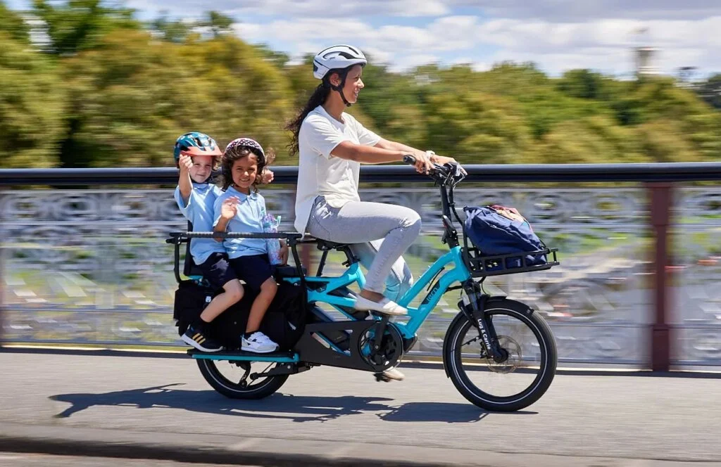 The Tern GSD ebike is a compact and versatile cargo bike designed to carry two passengers or children. The image shows the bike parked on a city street with a blurred cityscape in the background. The bike has a sturdy frame and a rear cargo rack, which is equipped with a comfortable and secure child seat. The child seat can be easily removed when not in use, and the cargo rack can accommodate other cargo, such as groceries or work equipment. The bike also features an electric motor and a battery, which provides assistance while riding, making it easier to navigate hills or long distances. The Tern GSD ebike is a perfect solution for families or commuters who need a compact and versatile bike that can carry multiple passengers or cargo. It is available at Lug+Carrie.