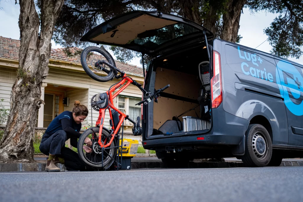 An energetic woman smiles as she effortlessly maneuvers an electric-powered Lug+Carrie ebike outside the vibrant backdrop of the Lug+Carrie van. Engaged in providing at-home services, she showcases the convenience and eco-friendliness of this modern transportation solution. Her confident posture and the seamless integration of the ebike with the van symbolize efficiency, versatility, and a commitment to sustainable practices.